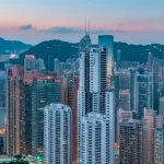 Immagine fotorealistica di un iconico edificio industriale di Hong Kong, riqualificato e integrato nel moderno skyline della città. Obiettivo grandangolare 20mm, fotografia architettonica al tramonto con lunga esposizione per enfatizzare le luci della città e la maestosità della struttura, dettagli nitidi.