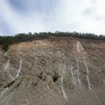 Wide-angle, 10-24mm, long exposure times, sharp focus: una vasta scarpata di una miniera a cielo aperto con evidenti segni di consolidamento tramite iniezione di resine poliuretaniche nelle fessure, sotto un cielo parzialmente nuvoloso, evidenziando la stabilità del pendio dopo l'intervento di rinforzo.