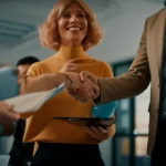 Una persona sorridente stringe la mano a un collega in un ambiente di lavoro luminoso e accogliente. Obiettivo prime 35mm, profondità di campo che sfoca leggermente lo sfondo, colori caldi e naturali, stile fotografico realistico.