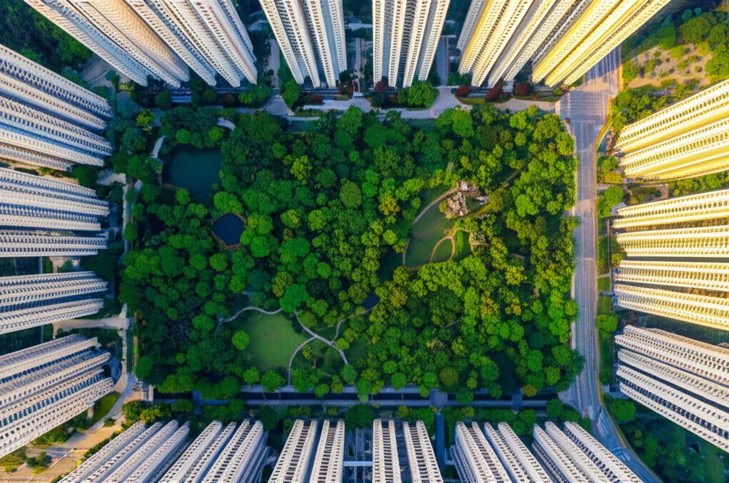 Fotografia aerea con drone, obiettivo grandangolare 18mm, di un grande parco urbano tropicale (come Bishan-Ang Mo Kio a Singapore) incastonato tra alti edifici residenziali. Si vede chiaramente il confine tra il verde lussureggiante del parco e il cemento circostante. Luce del tardo pomeriggio che crea ombre lunghe dagli alberi, mettendo in risalto la frescura del parco. Focus nitido, colori vividi.