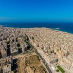 Immagine aerea grandangolare di Derna devastata dall'inondazione, con il percorso del fiume in piena che taglia la città e sfocia nel Mediterraneo. Wide-angle, 18mm, long exposure per acqua mossa, focus nitido sulla distruzione, colori desaturati per un effetto drammatico.