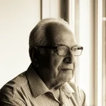 Ritratto fotografico di un uomo anziano sorridente e in salute, con occhiali, che guarda pensieroso fuori da una finestra. Luce naturale soffusa, prime lens 35mm, profondità di campo per mettere a fuoco il soggetto e sfocare leggermente lo sfondo, bianco e nero con un leggero viraggio seppia per un tocco classico e riflessivo.