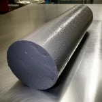 Un campione cilindrico di calcestruzzo geopolimerico grigio scuro appena sformato, con gocce d'acqua sulla superficie, appoggiato su un banco da laboratorio metallico. Luce laterale che ne esalta la texture porosa. Obiettivo macro 105mm, messa a fuoco precisa sui dettagli superficiali.