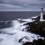 Grande angolo, una drammatica scena costiera durante un'ondata di tempesta con onde potenti illuminate da un faro lontano, lente da 14 mm, una lunga esposizione per il movimento dell'acqua liscio ma onde affilate, catturando la potenza grezza della natura e la necessità di previsioni accurate.
