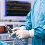Primo piano delle mani guantate di un anestesista che regolano con precisione una manopola su un'apparecchiatura di anestesia complessa in una sala operatoria moderna e ben illuminata. Sullo sfondo, sfocato, si intravede un monitor con tracciati vitali. Obiettivo macro, focale 90mm, illuminazione controllata tipica della sala operatoria, alta definizione dei dettagli sulle dita e sull'apparecchiatura.