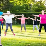 Un gruppo di donne di mezza età, diverse tra loro, che partecipano con entusiasmo a una lezione di ginnastica dolce in un parco cittadino, sotto la guida di un'istruttrice. Alcune sorridono, altre sono concentrate. Telephoto zoom, 150mm, fast shutter speed per cogliere il movimento, luce naturale del tardo pomeriggio.