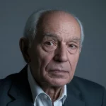 Ritratto fotografico di un uomo anziano (65-70 anni) dall'aspetto pensieroso ma speranzoso, scattato con obiettivo prime da 35mm. L'uomo guarda verso una fonte di luce laterale. Utilizzo di duotono blu e grigio per un'atmosfera seria ma non cupa. Profondità di campo ridotta (Depth of field) per sfocare leggermente lo sfondo, che suggerisce un ambiente medico moderno ma non invadente.