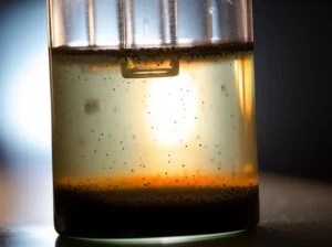Immagine fotorealistica di un reattore fotocatalitico in vetro illuminato da una luce intensa che simula il sole. All'interno, acqua leggermente torbida che si sta schiarendo, con nanoparticelle scure (il nanocomposito) sospese. Dettagli: obiettivo macro 60mm, alta definizione, illuminazione controllata per enfatizzare la trasparenza e le particelle, goccioline di condensa sul vetro.