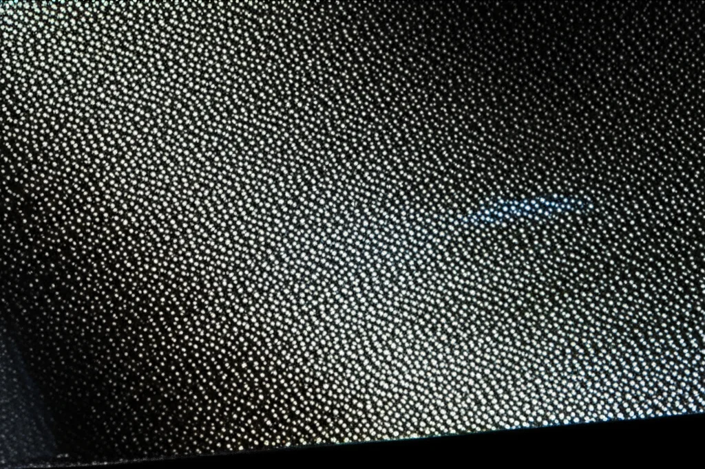 Primo piano di un film sottile di carbonio amorfo e nichel depositato su un substrato di vetro, con un raggio di luce che ne colpisce la superficie evidenziandone la micro-rugosità e le nanoparticelle. Obiettivo macro 100mm, illuminazione laterale controllata per esaltare i dettagli superficiali, alta definizione.