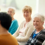 Un gruppo diversificato di persone anziane, uomini e donne, che sorridono e interagiscono positivamente durante una sessione informativa sulla salute in un ambiente luminoso e accogliente. Fotografia di gruppo, obiettivo 35mm, luce naturale calda, profondità di campo che mette a fuoco alcuni volti espressivi mentre altri sono leggermente sfocati sullo sfondo, trasmettendo un senso di comunità e coinvolgimento.
