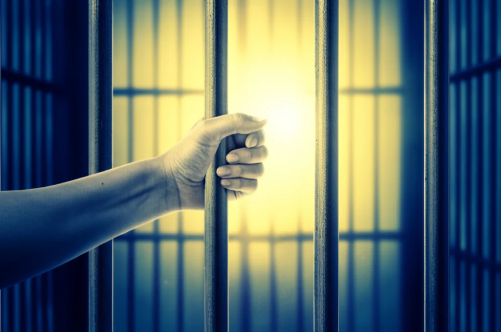 Conceptual image representing mental health support in a challenging environment: a guiding light or a helping hand reaching through prison bars. Prime lens, 35mm, duotone (e.g., warm yellow and cool blue), depth of field, symbolic.