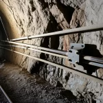 Immagine fotorealistica di una sezione di tunnel in roccia tenere, illuminata da luci di cantiere. In primo piano, un sistema di ancoraggio precompresso con una piastra d'ancoraggio ben visibile, installata contro la superficie irregolare della roccia. Dettagli: Prime lens, 35mm, profondità di campo, illuminazione drammatica per evidenziare la texture della roccia e i componenti metallici dell'ancoraggio.