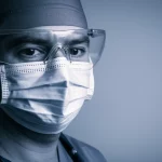 Ritratto intenso di un medico in prima linea durante la pandemia COVID-19, con evidenti segni di stanchezza ma sguardo determinato, indossa mascherina e visiera protettiva. L'illuminazione è drammatica, laterale, per enfatizzare l'espressione. Portrait photography, prime lens, 35mm, duotone blu freddo e grigio neutro, depth of field per isolare il soggetto.