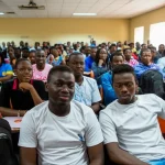 Un gruppo diversificato di studenti di medicina e infermieristica nigeriani, alcuni sorridenti altri pensierosi, in un'aula universitaria moderna ma con segni di risorse limitate. Fotografia di gruppo, obiettivo 35mm, luce naturale mista ad artificiale, per un'immagine realistica che catturi speranza e preoccupazione.