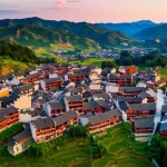 Fotografia aerea di un cluster di antichi villaggi cinesi annidati tra colline verdi e campi coltivati, al tramonto. Le case tradizionali con tetti di tegole scure creano un pattern armonioso. Obiettivo grandangolare 24mm, lunga esposizione per cieli soffusi, messa a fuoco nitida sul paesaggio.