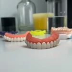 Fotografia still life di diverse basi per protesi dentali stampate in 3D e una convenzionale, alcune lucidate, altre vetrificate, disposte su un tavolo da laboratorio dentale pulito. Luce da studio controllata, lente macro 90mm, alta definizione, focus preciso sulle differenze superficiali.