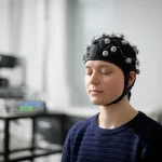 A photorealistic 35mm portrait of a young adult participant wearing an fNIRS cap with probes, sitting calmly in a quiet, softly lit room, eyes closed, with a subtle depth of field blurring the background equipment. High detail, controlled lighting.
