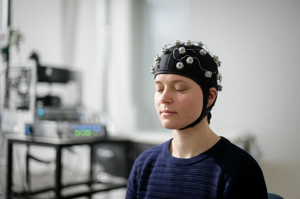 A photorealistic 35mm portrait of a young adult participant wearing an fNIRS cap with probes, sitting calmly in a quiet, softly lit room, eyes closed, with a subtle depth of field blurring the background equipment. High detail, controlled lighting.