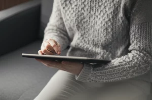 Una persona seduta comodamente su un divano che utilizza un tablet per cercare informazioni sulla salute. La luce naturale illumina la scena. Obiettivo prime, 35mm, profondità di campo che mette a fuoco il tablet e il volto concentrato della persona, atmosfera rilassata e domestica.