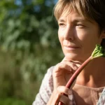 A woman in her early 50s looking serene and thoughtful, holding a small sprig of rhubarb. Soft natural light illuminates her face. 35mm portrait lens, depth of field, creating a gentle, hopeful mood.