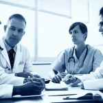 Fotografia di un team medico multidisciplinare etiope (oncologo, infermiera, ginecologo) in una sala riunioni di un ospedale, discutono seriamente attorno a un tavolo con documenti sparsi, luce soffusa da una finestra laterale, obiettivo da 24mm per includere il gruppo, toni blu e grigio duotone, profondità di campo media per mantenere a fuoco i volti e i documenti.