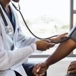 Immagine di un medico che misura la pressione sanguigna a un paziente in un ambiente clinico moderno e luminoso a Città del Capo. Obiettivo da 35mm, profondità di campo, colori naturali, per illustrare il tema della diagnosi e cura dell'ipertensione.