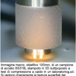 Immagine macro, obiettivo 105mm, di un campione di acciaio SS316L stampato in 3D sottoposto a test di compressione a caldo in un laboratorio. Si vedono chiaramente le texture superficiali del metallo, con zone incandescenti a causa dell'alta temperatura. Illuminazione controllata e precisa messa a fuoco sui dettagli della deformazione.