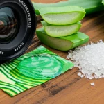 Macro lens, 105mm, un campione di tessuto ecologico stampato con motivi vivaci realizzati con biomateriali, esposto accanto agli ingredienti grezzi come gel di aloe vera trasparente e scaglie di chitosano biancastre, il tutto su una superficie di legno rustico, alta definizione, illuminazione da studio controllata per esaltare texture e colori.