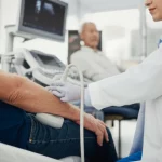 Primo piano di un medico che esegue un'ecografia addominale su un paziente anziano in un ambiente clinico luminoso. Lente prime 35mm, profondità di campo, colori naturali, per trasmettere cura e tecnologia avanzata nella diagnosi della fragilità e sarcopenia.