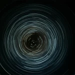 Concettualizzazione astratta delle oscillazioni chirali di una particella di Dirac, con scie luminose che rappresentano il cambiamento di chiralità nel tempo. Wide-angle, 10mm, long exposure, smooth light trails, sharp focus on a central particle representation, con un orologio stilizzato sullo sfondo a indicare il tempo finito.