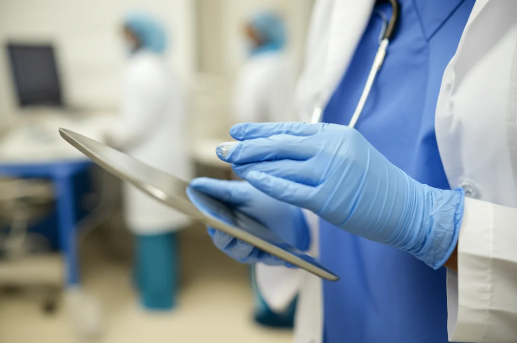 Primo piano delle mani guantate di un'infermiera etiope che consulta linee guida mediche su un tablet moderno, con lo sfondo sfocato di un pronto soccorso etiope attivo ma ordinato. Macro lens 85mm, high detail, luce laterale morbida che illumina le mani e il tablet.