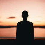Una silhouette di una persona che guarda un orizzonte luminoso e speranzoso da una stanza più scura, simboleggiando la lotta contro la malattia (cancro) e la depressione. Fotografia wide-angle 24mm, colori caldi e vibranti all'orizzonte (alba/tramonto) contrastanti con toni freddi e ombre all'interno della stanza, focus nitido sull'orizzonte lontano.