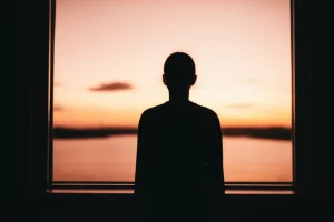 Una silhouette di una persona che guarda un orizzonte luminoso e speranzoso da una stanza più scura, simboleggiando la lotta contro la malattia (cancro) e la depressione. Fotografia wide-angle 24mm, colori caldi e vibranti all'orizzonte (alba/tramonto) contrastanti con toni freddi e ombre all'interno della stanza, focus nitido sull'orizzonte lontano.