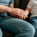 Fotografia realistica a colori, obiettivo 50mm, luce naturale morbida da una finestra. Un genitore (viso fuori campo o sfocato per privacy) tiene dolcemente la mano di un bambino seduto accanto a lui/lei su un divano. L'atmosfera è calma e intima, suggerendo cura e supporto nel contesto dell'epilessia infantile. Profondità di campo media.