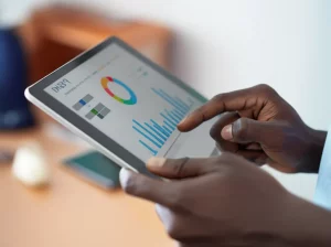 Primo piano fotorealistico delle mani di un operatore sanitario africano che digitano attentamente su un tablet, visualizzando grafici e dati nutrizionali del sistema DHIS2. L'ambientazione è un centro sanitario rurale luminoso in Burkina Faso. Obiettivo macro 85mm, illuminazione naturale controllata, alta definizione dei dettagli dello schermo, delle mani e della texture della divisa, profondità di campo ridotta per isolare il soggetto.