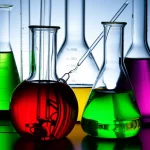 Fotografia macro di una reazione chimica in corso, 100mm, alta definizione, messa a fuoco precisa, illuminazione controllata. Vetreria da laboratorio contenente soluzioni colorate e brillanti, con un accenno a catalizzatori metallici (nichel), che simboleggia la sintesi di esteri benzil boronici.