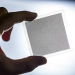 Immagine fotorealistica, obiettivo prime 35mm, che mostra un primo piano di una mano che tiene un foglio flessibile e quasi invisibile (la metasuperficie) contro uno sfondo luminoso, dimostrandone la trasparenza e la flessibilità. Sottili pattern geometrici (simili all'ITO) luccicano sulla superficie. Effetto di profondità di campo che sfoca leggermente lo sfondo.