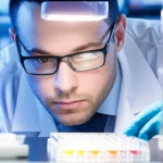 Ricercatore in camice bianco che osserva attentamente una piastra multi-pozzetto contenente campioni biologici sotto una luce da laboratorio, sfondo sfocato di attrezzature scientifiche, fotografia ritratto 35mm, profondità di campo.