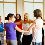 Un gruppo eterogeneo di donne sorridenti che ballano insieme in cerchio durante un vivace corso di danza comunitaria, simboleggiando l'inclusione e la gioia. Obiettivo grandangolare, 24mm, colori caldi e luminosi, catturando il movimento e la connessione tra le partecipanti in un ambiente accogliente.