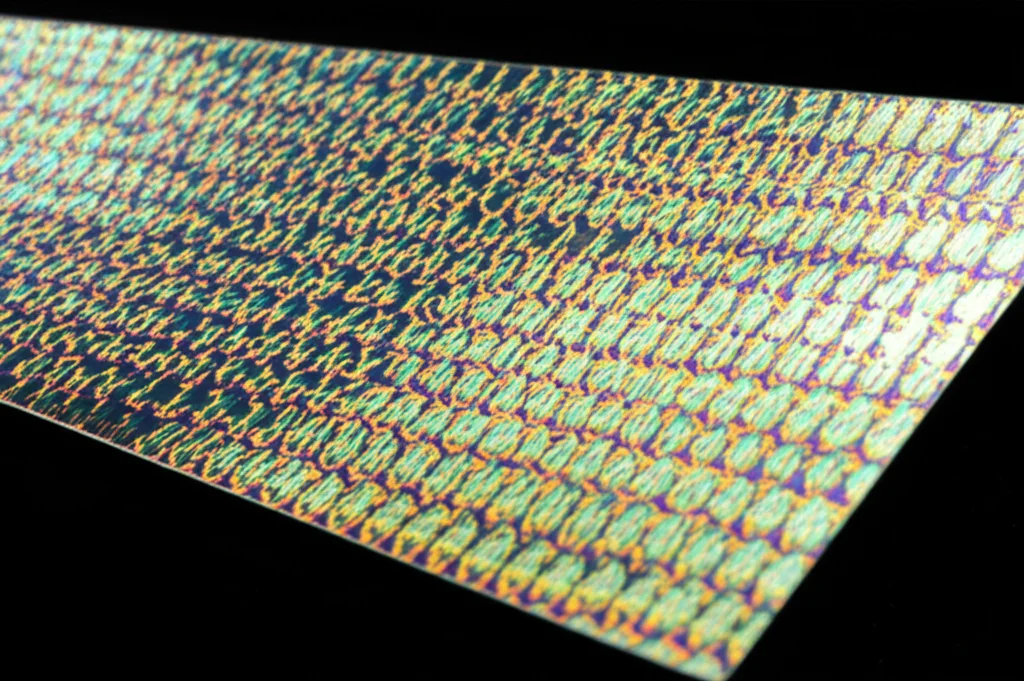 Immagine fotorealistica di un film sottile di perovskite, leggermente iridescente, illuminato da un fascio di luce focalizzato, che evidenzia l'interazione tra luce e struttura del materiale, obiettivo macro 70mm, alto dettaglio, illuminazione controllata.