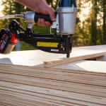 A person using a pneumatic nail gun on stacked lumber boards, creating a mass timber panel, with a sun-dappled California forest visible in the background. Macro lens, 60mm, high detail, controlled lighting.