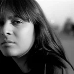 Portrait photography che cattura la resilienza e la vulnerabilità di una giovane donna transgender a Lima, Perù. Obiettivo 35mm prime, luce naturale morbida, profondità di campo ridotta che sfoca leggermente lo sfondo urbano, stile film noir che enfatizza ombre e luci, trasmettendo introspezione e forza in mezzo alle avversità.