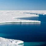 Fotografia paesaggistica artica, obiettivo grandangolare 15mm, che mostra la transizione tra una vasta distesa di ghiaccio marino bianco e l'acqua oceanica aperta di colore blu intenso sotto un cielo limpido. Luce solare radente che crea lunghe ombre sul ghiaccio. Messa a fuoco nitida sull'orizzonte, lunga esposizione per rendere l'acqua leggermente setosa.
