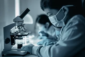 Un gruppo eterogeneo di bambini di diverse età, dai neonati agli adolescenti, che simboleggiano le diverse fasce d'età colpite dal COVID-19, osservati attraverso la lente di un microscopio stilizzato. Fotografia concettuale, obiettivo da 35mm, luce soffusa e drammatica con toni blu e grigi duotone, profondità di campo che mette a fuoco alcuni bambini mentre altri sono leggermente sfocati per rappresentare le diverse ondate e impatti.