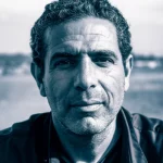 Un ritratto intenso di un veterano israeliano, il suo volto segnato dall'esperienza ma con uno sguardo che cerca integrazione. L'immagine utilizza una lente prime da 35mm, con un effetto duotone blu e grigio per enfatizzare l'introspezione. Profondità di campo ridotta per isolare il soggetto. Stile fotorealistico.