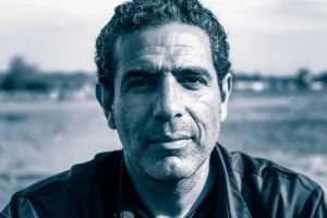 Un ritratto intenso di un veterano israeliano, il suo volto segnato dall'esperienza ma con uno sguardo che cerca integrazione. L'immagine utilizza una lente prime da 35mm, con un effetto duotone blu e grigio per enfatizzare l'introspezione. Profondità di campo ridotta per isolare il soggetto. Stile fotorealistico.