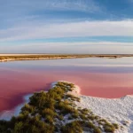 Paesaggio grandangolare di un lago salato dai colori surreali nella regione di Dingbian, Cina, con evidenti depositi di sale cristallizzato bianco e rosa sulle rive e una scarsa vegetazione alofita di colore verde scuro. Luce del tardo pomeriggio che crea lunghe ombre e accentua le texture del terreno salino e screpolato. Obiettivo grandangolare 10-24mm, f/11, messa a fuoco nitida su tutto il campo, lunga esposizione per rendere l'acqua del lago setosa e riflettente il cielo parzialmente nuvoloso.