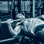 Portrait photography di un uomo giovane e muscoloso, in una palestra moderna, mentre esegue un esercizio di panca piana con bilanciere. L'immagine è scattata con un obiettivo da 35mm, con profondità di campo che mette a fuoco l'atleta e sfoca leggermente lo sfondo. Dettagli nitidi sui muscoli pettorali e tricipiti in contrazione. Duotone blu e grigio per un effetto intenso.