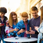 Un gruppo diversificato di adolescenti che interagiscono con i loro smartphone in un ambiente sociale informale, come una caffetteria all'aperto o un parco. Alcuni sorridono e chattano tra loro mostrando gli schermi, altri sono più concentrati individualmente sui loro dispositivi. L'immagine deve trasmettere sia l'aspetto sociale che quello individuale dell'uso dello smartphone. Obiettivo prime da 35mm, luce naturale dorata del tardo pomeriggio, profondità di campo media per mantenere a fuoco più soggetti, colori vivaci ma realistici, stile fotografia di reportage.