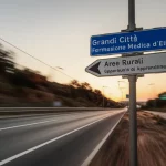 Un bivio stradale con un cartello che indica 'Grandi Città - Formazione Medica d'Elite' e un altro, più piccolo e meno curato, 'Aree Rurali - Opportunità di Apprendimento'. Telephoto zoom, 100mm, fast shutter speed per catturare un momento di scelta, luce del tardo pomeriggio che crea lunghe ombre e un'atmosfera di dilemma.